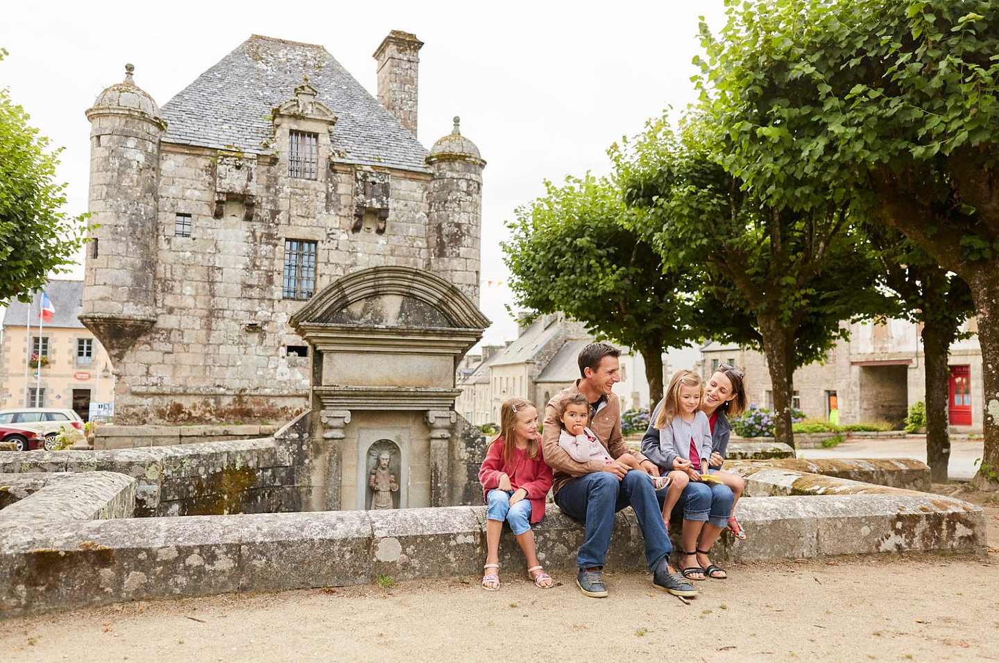 Famille heureuse devant le Pr&eacute;sidial de Guerlesquin, un b&acirc;timent historique en Bretagne, entour&eacute; de verdure et d\'architecture traditionnelle