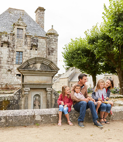 Famille heureuse devant le Pr&eacute;sidial de Guerlesquin, un b&acirc;timent historique en Bretagne, entour&eacute; de verdure et d\'architecture traditionnelle