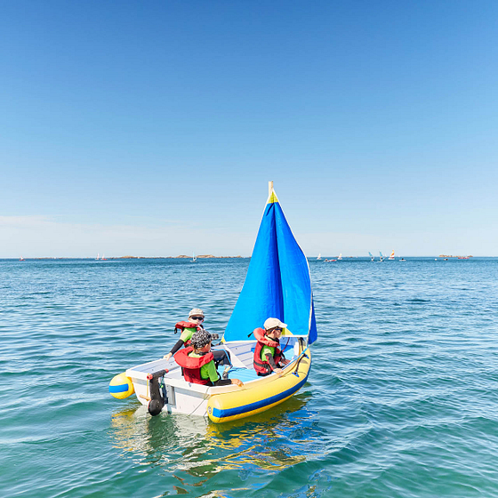 Trois enfants naviguant sur un petit voilier gonflable avec un drapeau bleu vif au large de Carantec.