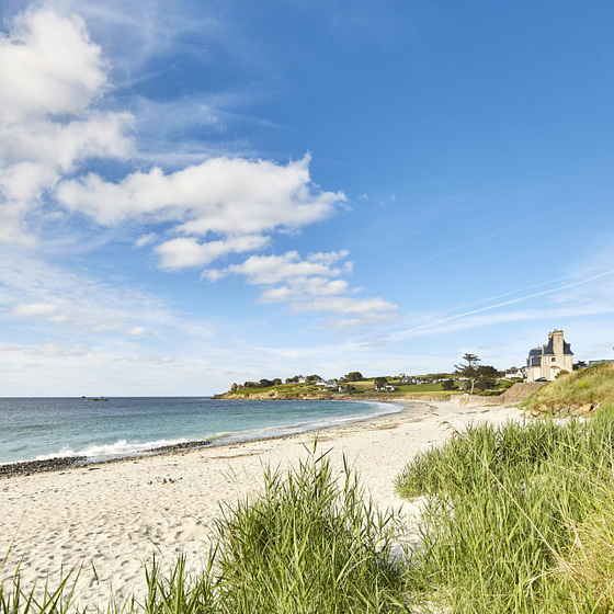 Plage de Saint-Samson &agrave; Plougasnou avec son sable fin, vue sur une maison en pierre bretonne typique et des dunes verdoyantes sous un ciel bleu.