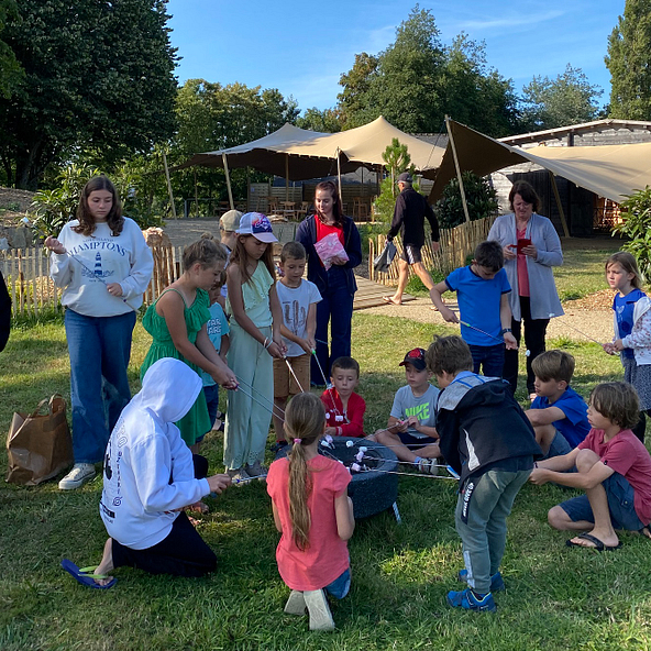 Des enfants réunis autour d\'un feu de camp improvisé grillant des chamallows au Domaine de Mesqueau, dans une ambiance joyeuse et animée.