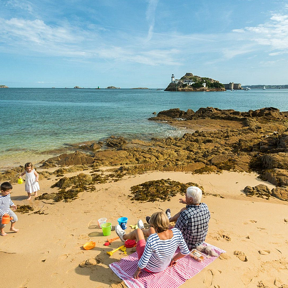 Famille profitant d&rsquo;un pique-nique sur une plage de Carantec, pr&egrave;s du camping Domaine de Mesqueau, avec vue sur l&rsquo;&icirc;le Lou&euml;t et le Ch&acirc;teau du Taureau.