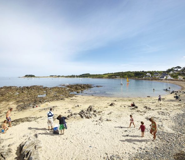 Plage de Térénez à Plougasnou, une crique animée avec des familles profitant du sable doré, des rochers en bordure, et des voiles colorées flottant sur l\'eau calme. En arrière-plan, des maisons typiques bretonnes et une végétation luxuriante.