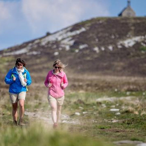 Deux randonneuses marchant sur un sentier dans les Monts d&rsquo;Arr&eacute;e, avec la chapelle Saint-Michel de Brasparts en arri&egrave;re-plan.
